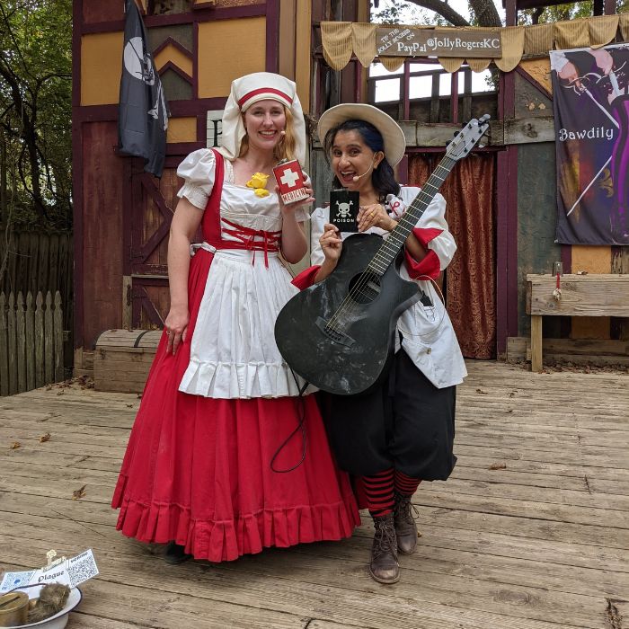 Lindsay and Manu. Renaissance Nurse in a big red dress with white apron and nurse cap. Medieval doctor in white lab coat with old style doctors hat and guitar. They both hold up medical flasks.