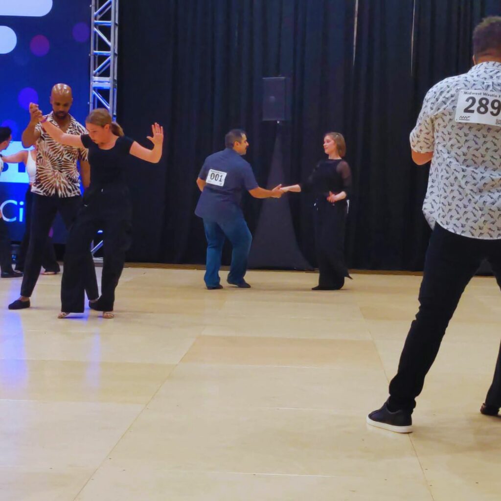 2 couples compete at Midwest Westie Fest in West Coast Swing Dance while a third couple lunges off the ride side of the photo.    Dance competition numbers are visible on backs.  