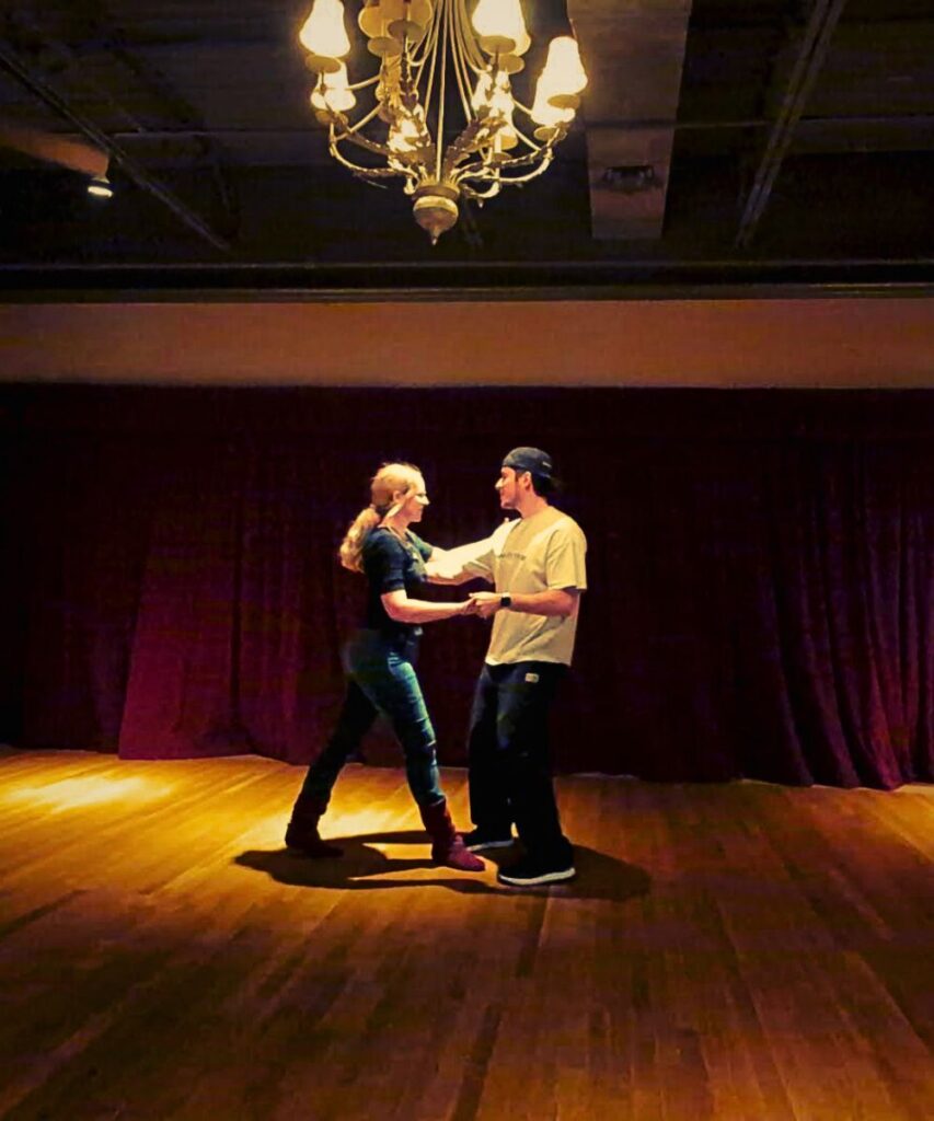 Lindsay West Coast Swing dancing in closed portion of whip with Instructor Jesse at CAST in front of red curtain on golden hardwood floor