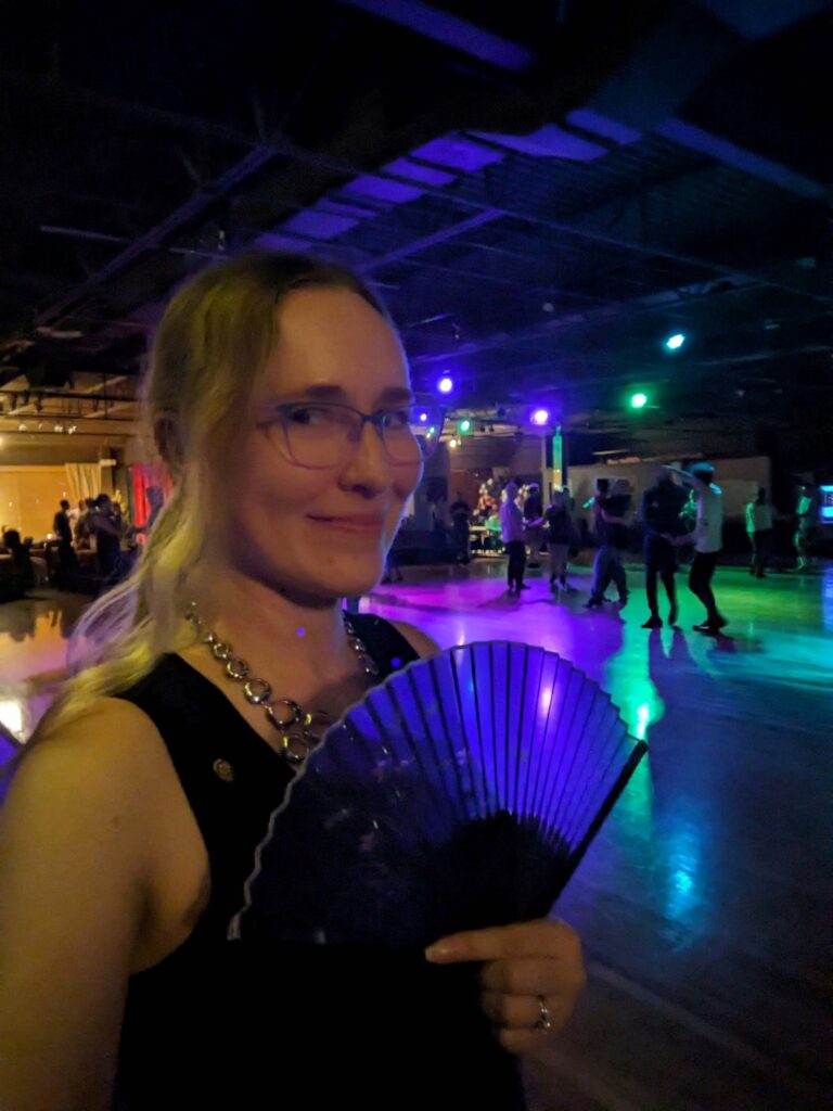 Lindsay holding a fan in front of West Coast Swing Dancers with Purple and Green lighting at CAST