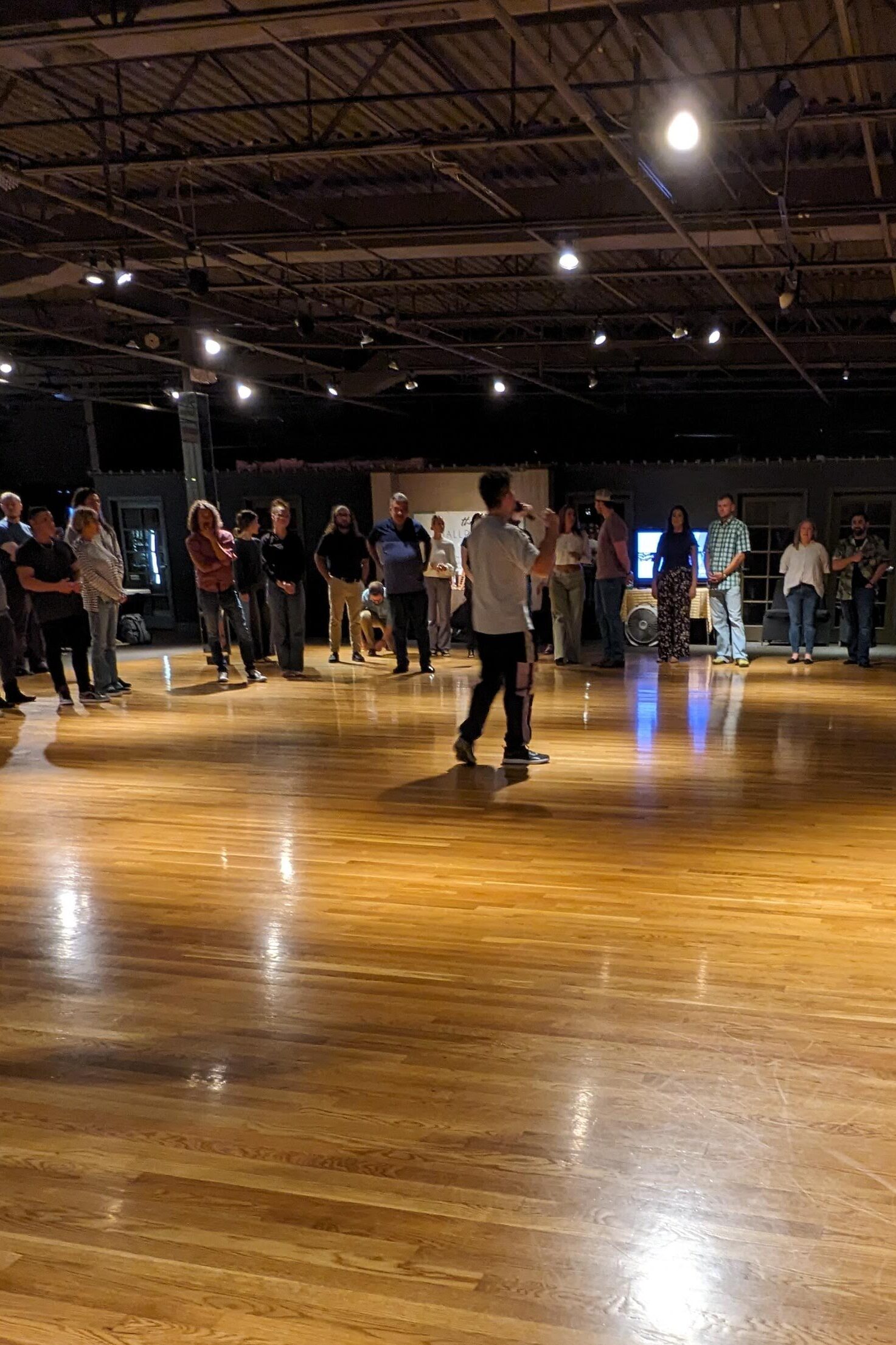 A large group of West Coast Swing Dance Students listen in a circle around the instructor on a golden ,wooden floor