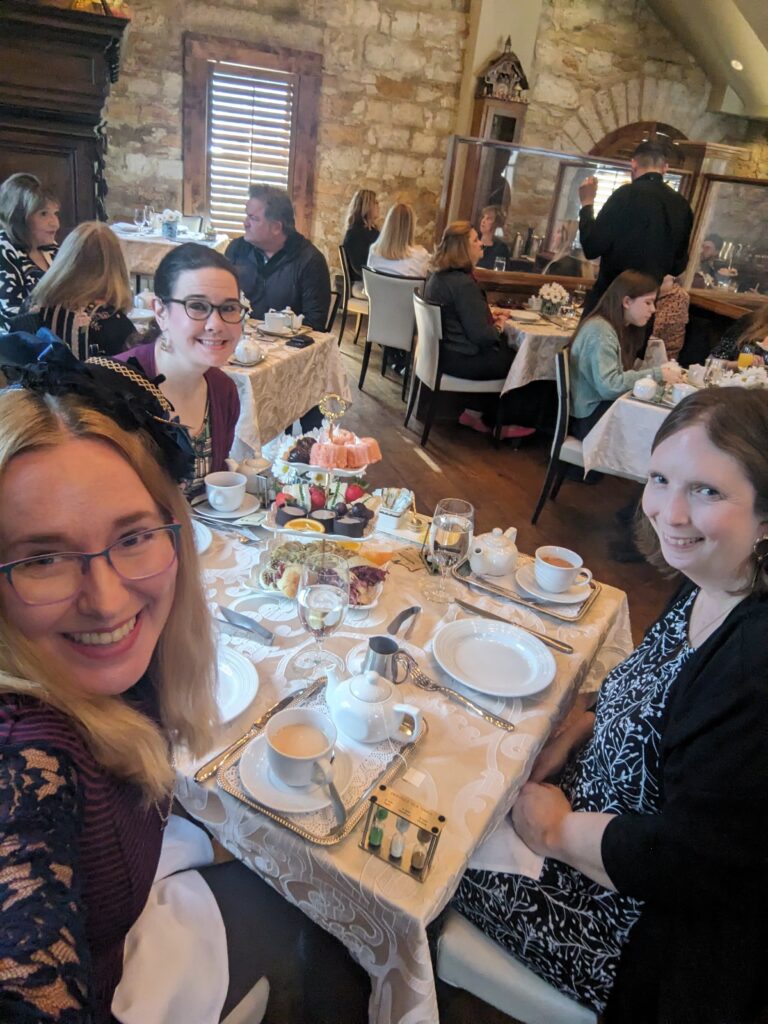 Lindsay and friends in tea finery sit around a table of tea treats in a Castle.