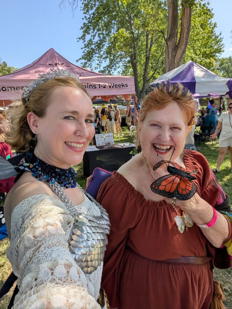 Lindsay in a white dress with chain mail top poses next to her mother with a butterfly puppet on her finger. Both smile at the Topeka Renfest.