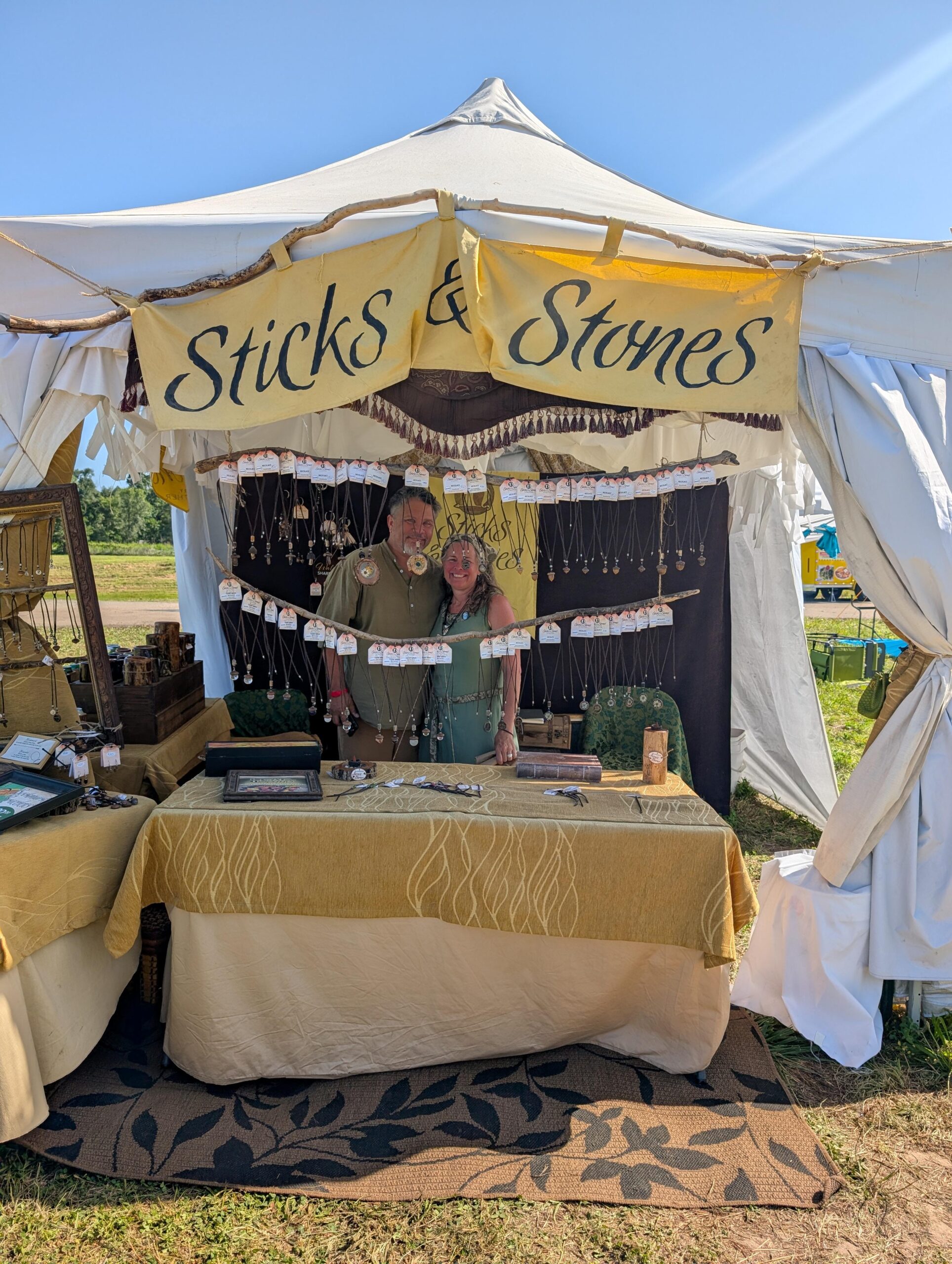 Vendor Tent at Topeka Renaissaince Festival. A couple stand proudly wit their wares under a sign reading Sticks and Stones with jewelry hanging along the front.