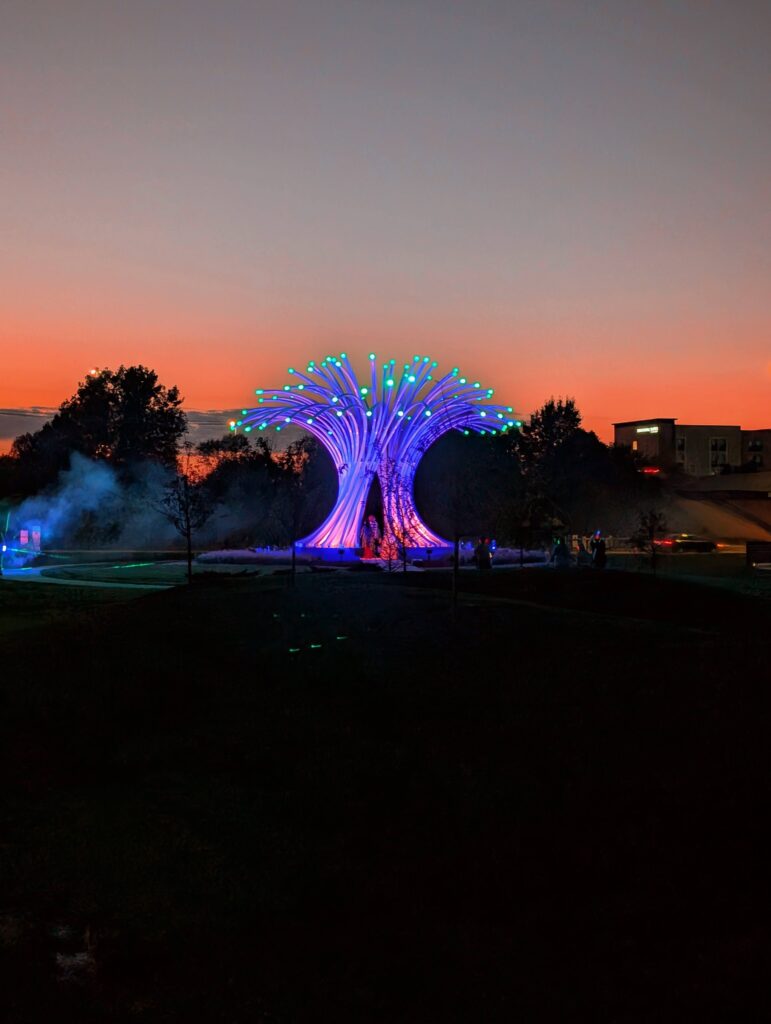 A purple modern sculpture with green lights on the ends is illuminated in front of a sunset. The sculpture looks like a tree or fountain made of a series of tubes.
